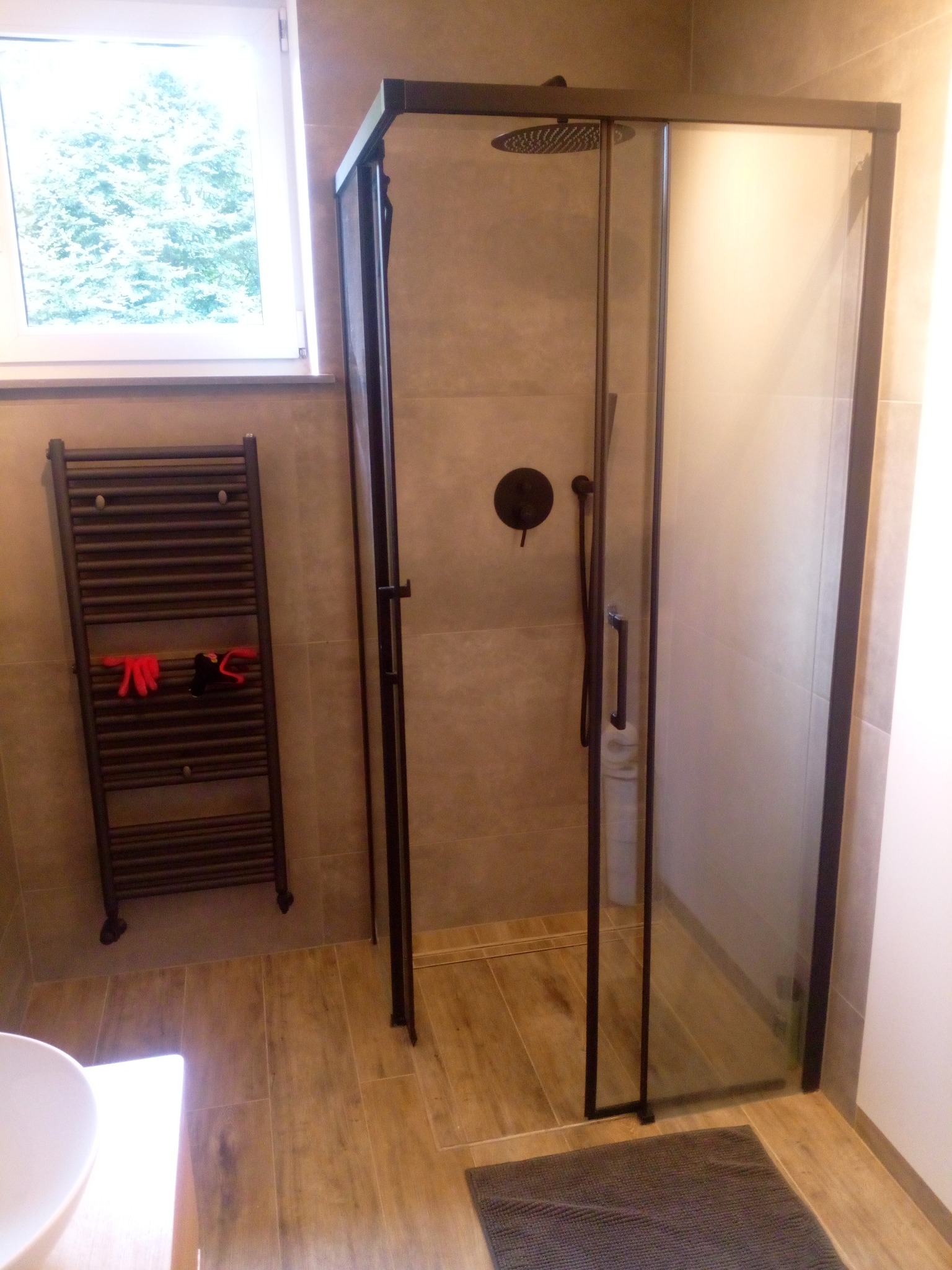 Nowoczesna kabina prysznicowa z czarną armaturą i grzejnikiem drabinkowym w wykończonej łazience. Jasne płytki imitujące drewno na podłodze, okno z widokiem na zieleń.