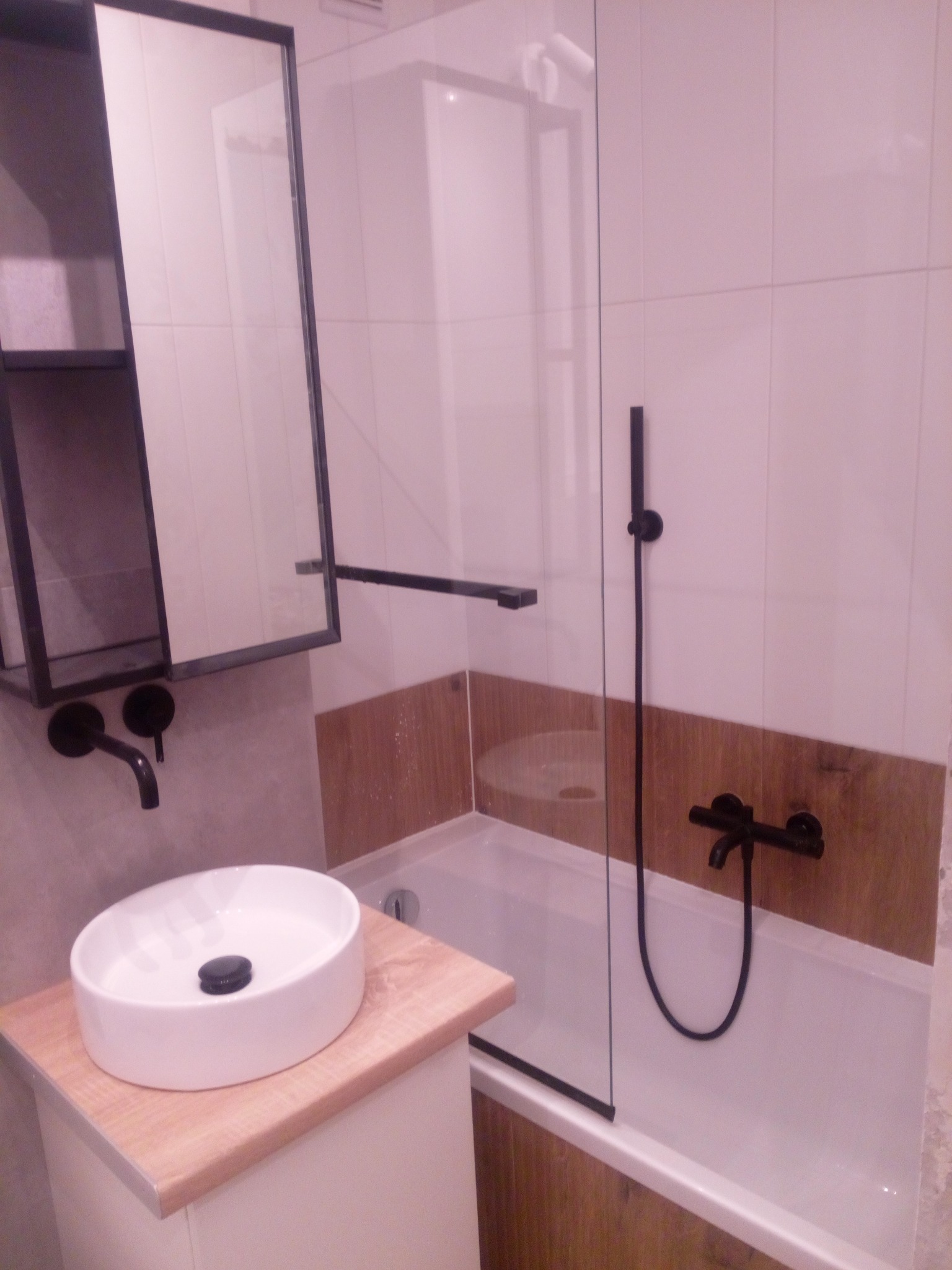 Nowoczesna łazienka z białą umywalką nablatową, czarną armaturą i kabiną prysznicową z elementami drewna. Minimalistyczny design i funkcjonalne rozwiązania.