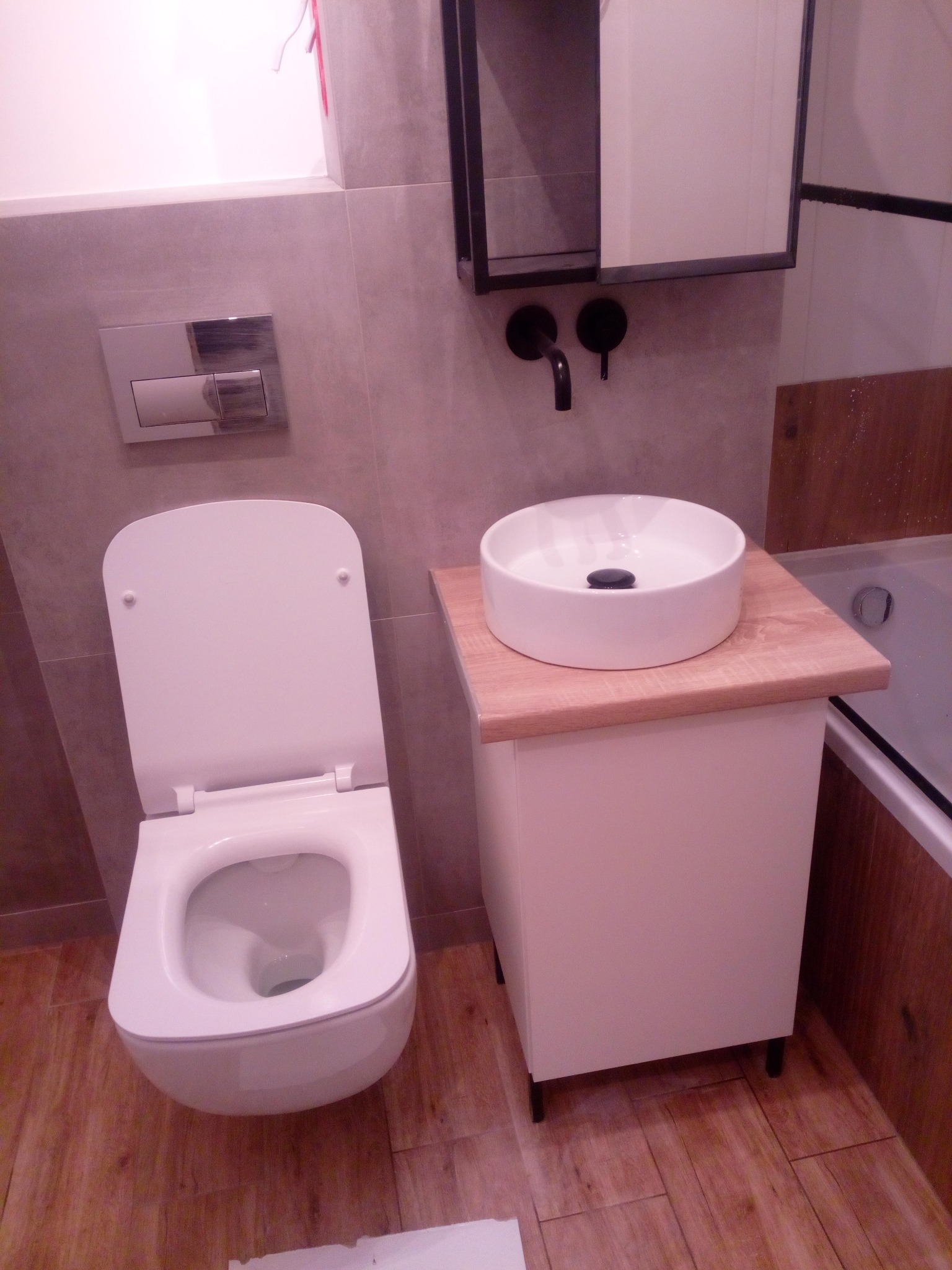 Nowoczesna łazienka: wisząca toaleta, okrągła umywalka nablatowa na szafce z drewnianym blatem, czarna armatura, płytki imitujące drewno na podłodze i ścianie.