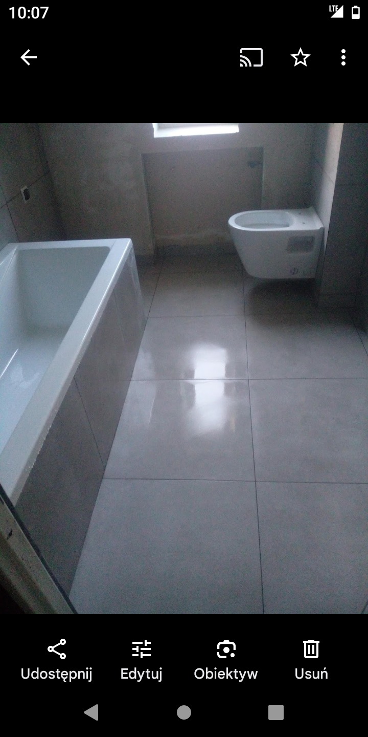 Wykończona łazienka z białą wanną, podwieszaną toaletą i szarymi płytkami podłogowymi, oświetlona naturalnym światłem z okna.