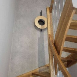 Pomalowany Wrocław - Drewniane schody ze stalowymi relingami przy szarej ścianie z dekoracyjnym, okrągłym elementem ściennym. Ujęcie z dołu, widok na fragment schodów i ścianę.