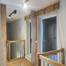 Pomalowany Wrocław - Nowoczesne wykończenie korytarza z lustrami, drewnianą balustradą i oknami z bambusowymi roletami. Oświetlenie punktowe podkreśla detale.