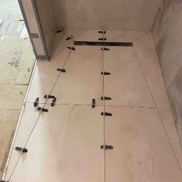 Pomalowany Wrocław - Układanie beżowych płytek podłogowych w łazience, z widocznym odpływem liniowym i czarnymi krzyżykami dystansowymi. Praca w toku, widoczne kartony po płytkach.