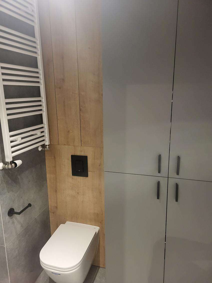 Nowoczesna łazienka: podwieszana toaleta z czarnym przyciskiem, grzejnik drabinkowy, szafka z szarymi frontami i drewnopodobne wykończenie ściany.