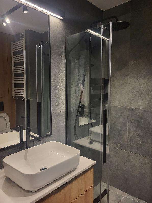 Nowoczesna łazienka z kabiną prysznicową typu walk-in, umywalką nablatową i szarymi płytkami imitującymi kamień, oświetlona listwą LED.