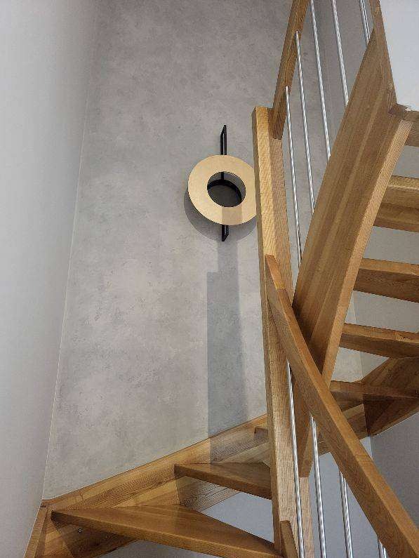 Drewniane schody ze stalowymi relingami przy szarej ścianie z dekoracyjnym, okrągłym elementem ściennym. Ujęcie z dołu, widok na fragment schodów i ścianę.