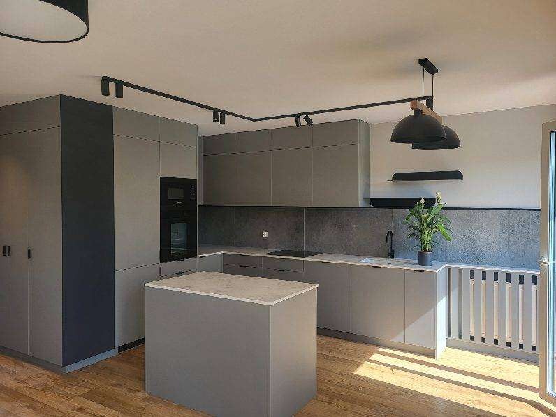 Nowoczesna szara kuchnia z wyspą, oświetleniem punktowym i drewnianą podłogą. Minimalistyczny design z czarnymi akcentami i rośliną doniczkową.