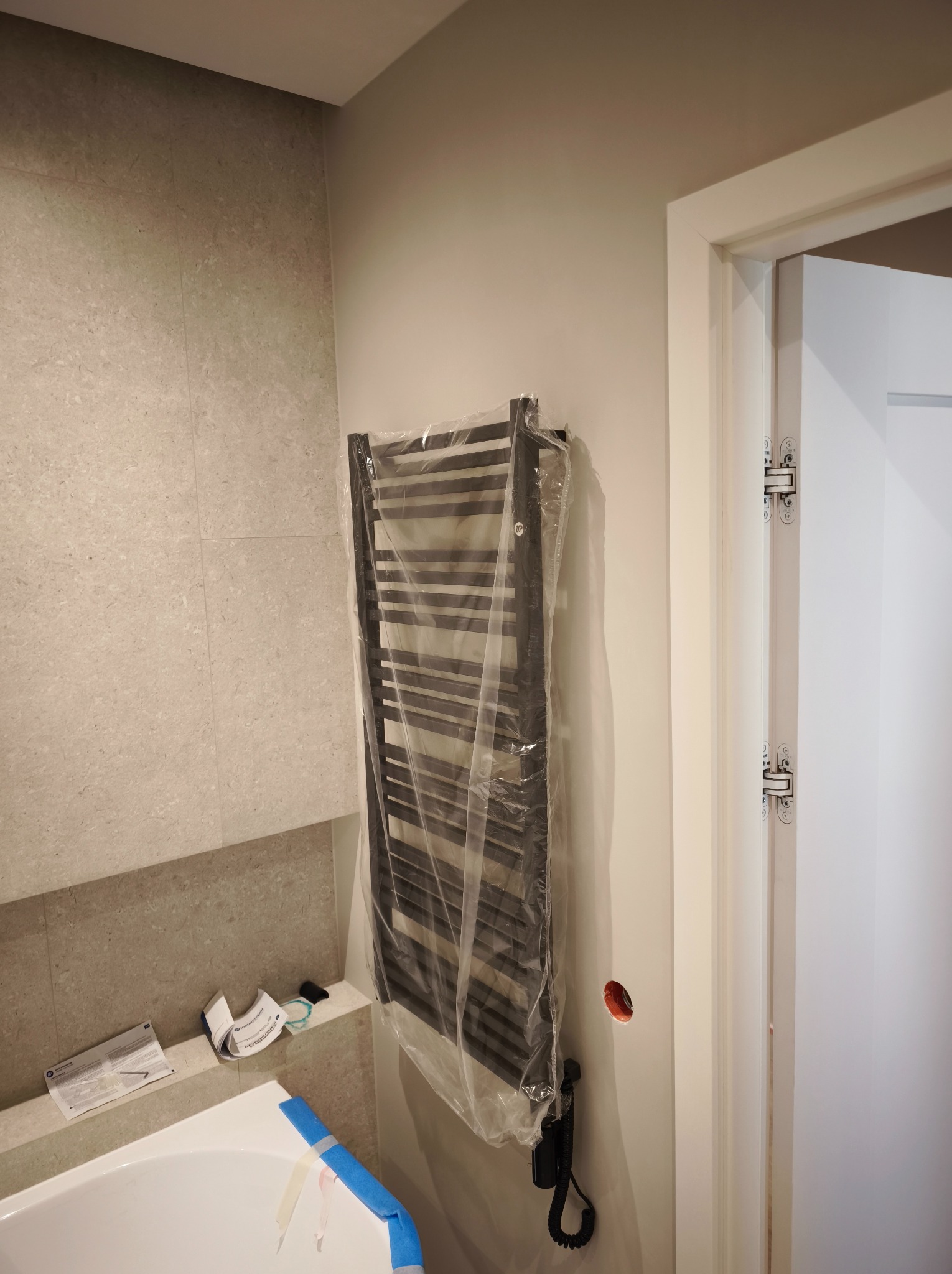 Nowy, czarny grzejnik łazienkowy, jeszcze w folii ochronnej, zamontowany na szarej ścianie z płytek. W tle widoczna biała wanna i fragment drzwi.