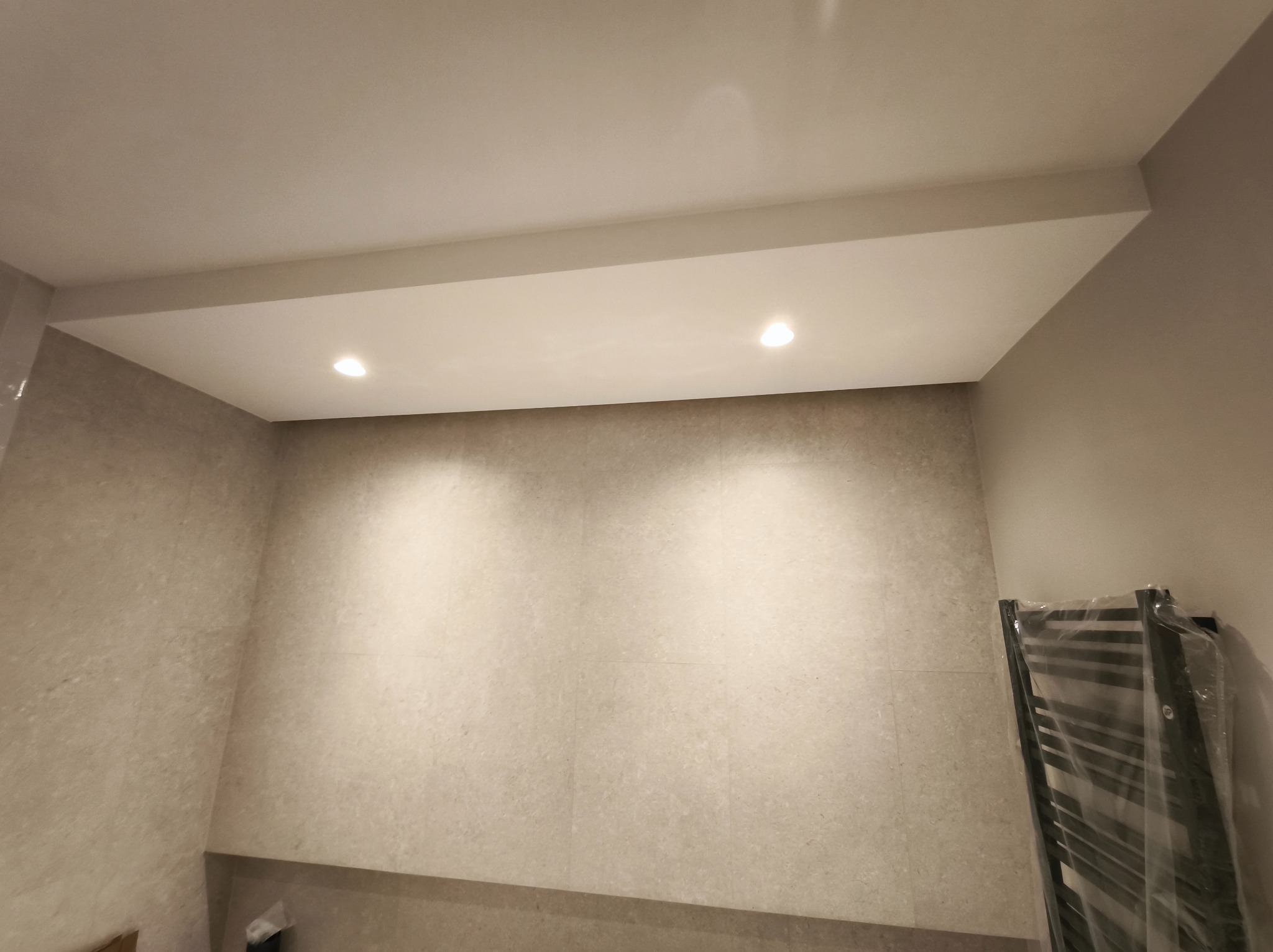 Wykończona łazienka z beżowymi płytkami, oświetleniem punktowym w suficie i grzejnikiem drabinkowym w folii ochronnej. Minimalistyczny design.