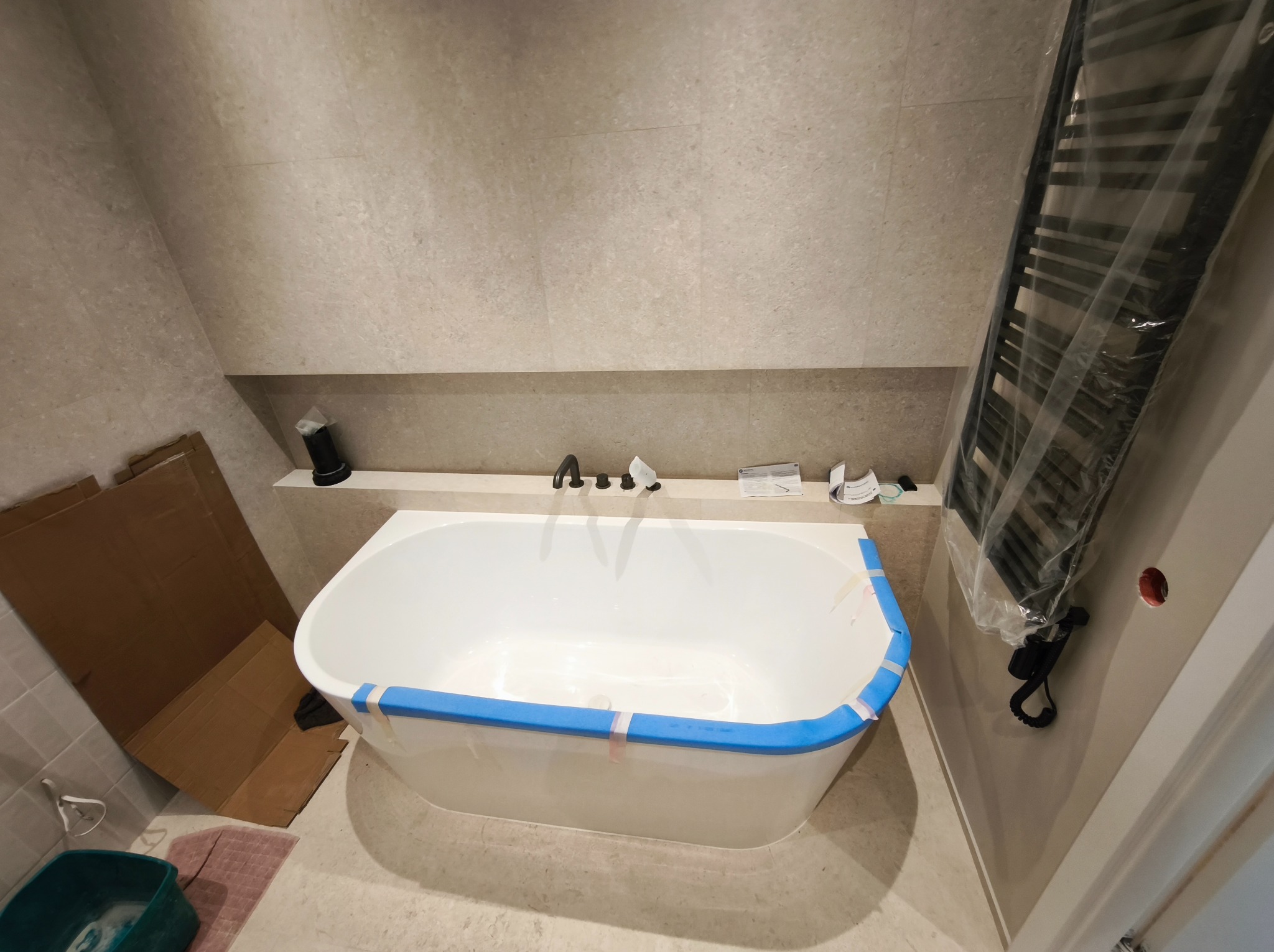 Biała wanna z niebieską taśmą zabezpieczającą w jasnej łazience, z grzejnikiem drabinkowym w folii i kartonami w rogu. Widoczne płytki w odcieniach beżu.