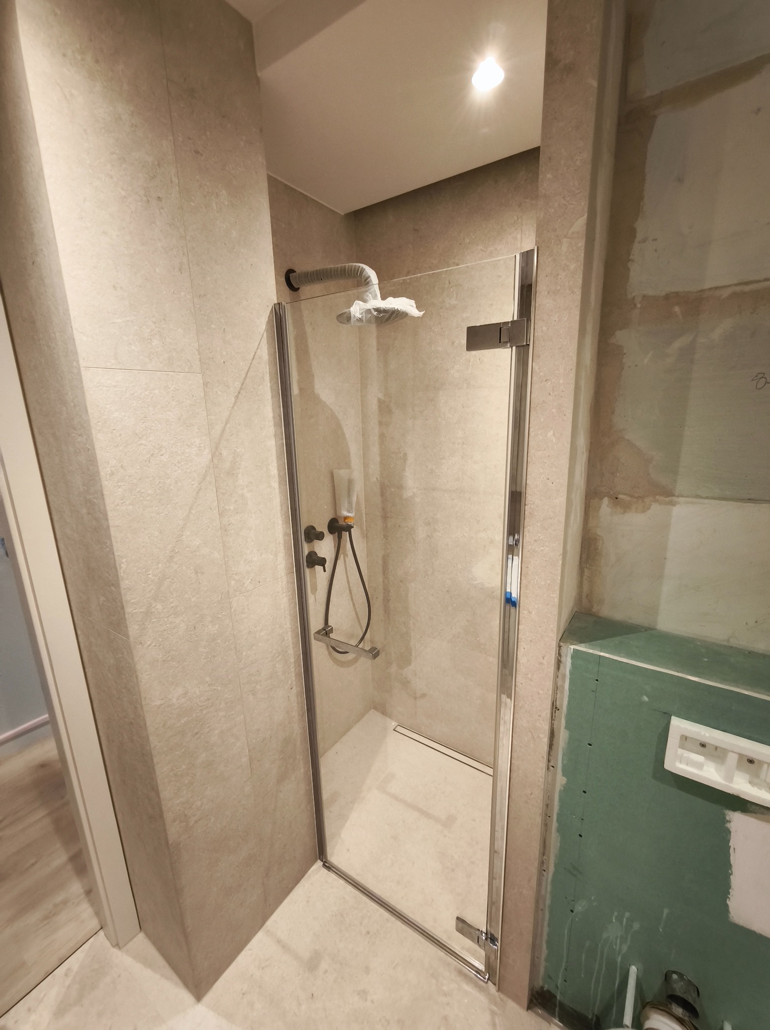 Kabina prysznicowa ze szklanymi drzwiami, jasne płytki na ścianach, widoczny fragment ściany z płytą gipsowo-kartonową. Nowoczesna armatura.