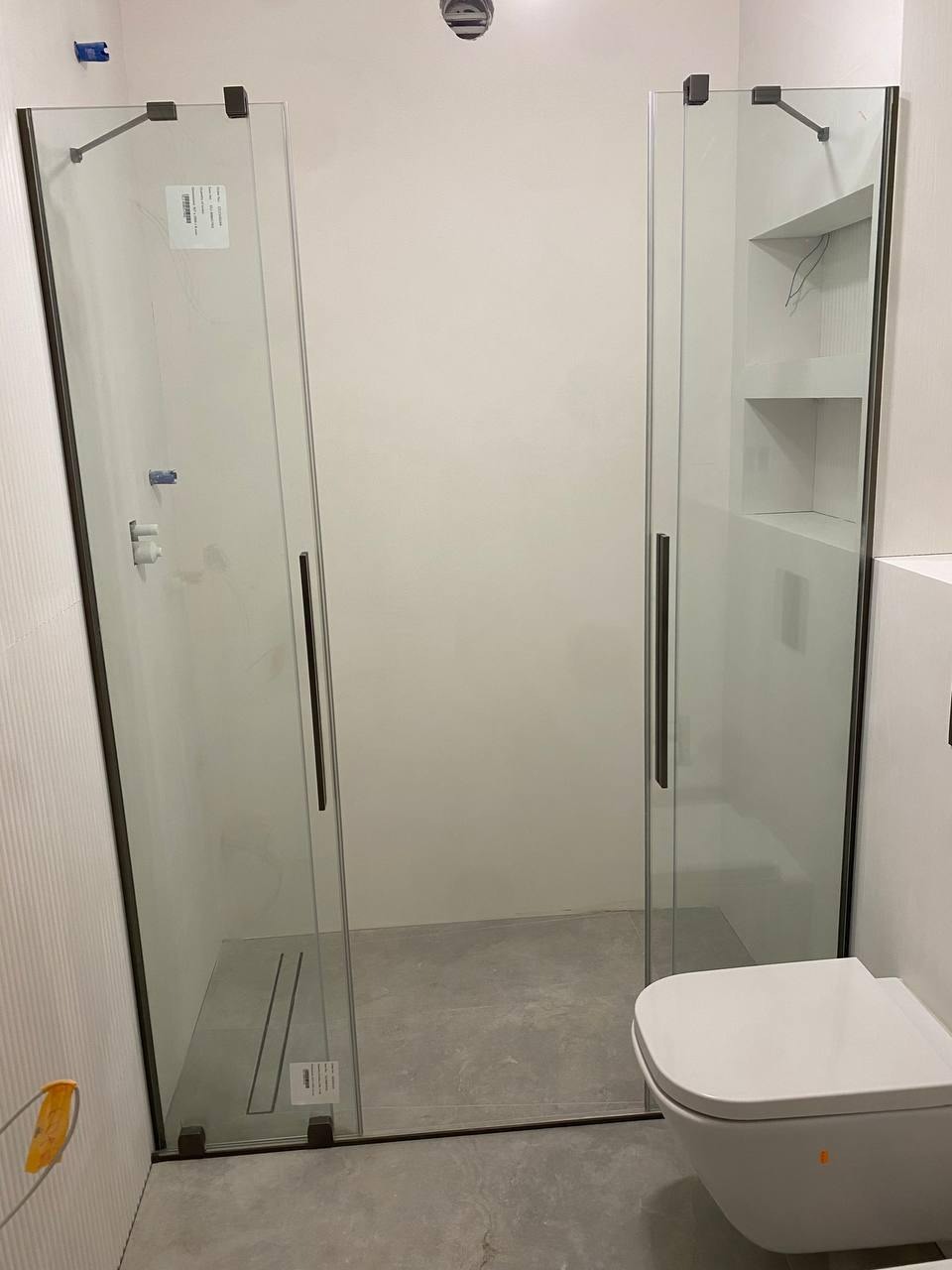 Nowoczesna kabina prysznicowa z grafitowymi okuciami i wnęką z półkami, minimalistyczny styl, jasna łazienka w trakcie wykańczania, widoczna toaleta podwieszana.