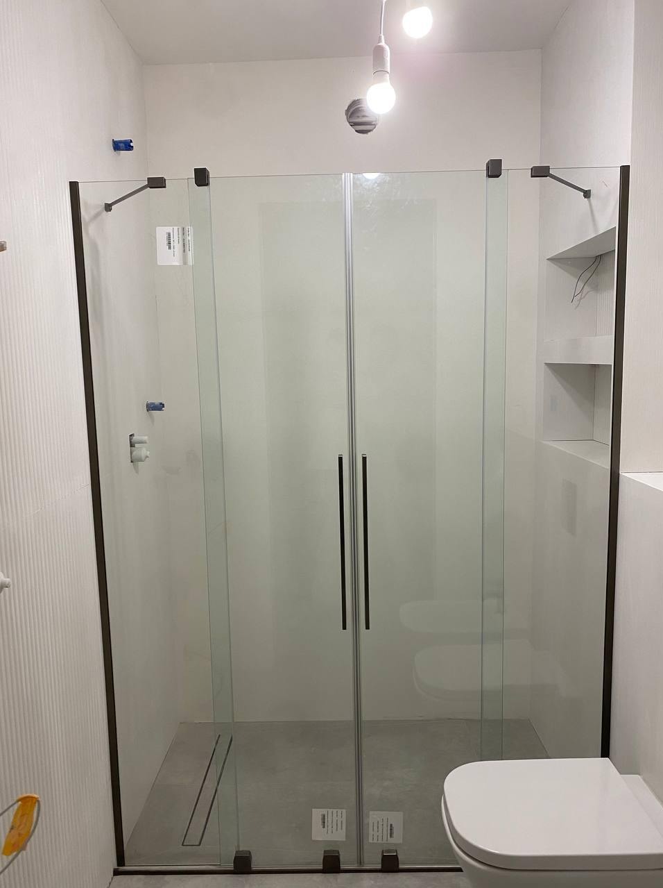 Nowoczesna kabina prysznicowa z czarnymi detalami w jasnej łazience. Widoczny odpływ liniowy i minimalistyczna armatura. Oświetlenie sufitowe podkreśla czystość wykończenia.