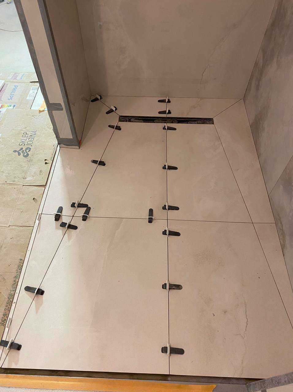 Układanie beżowych płytek podłogowych w łazience, z widocznym odpływem liniowym i czarnymi krzyżykami dystansowymi. Praca w toku, widoczne kartony po płytkach.