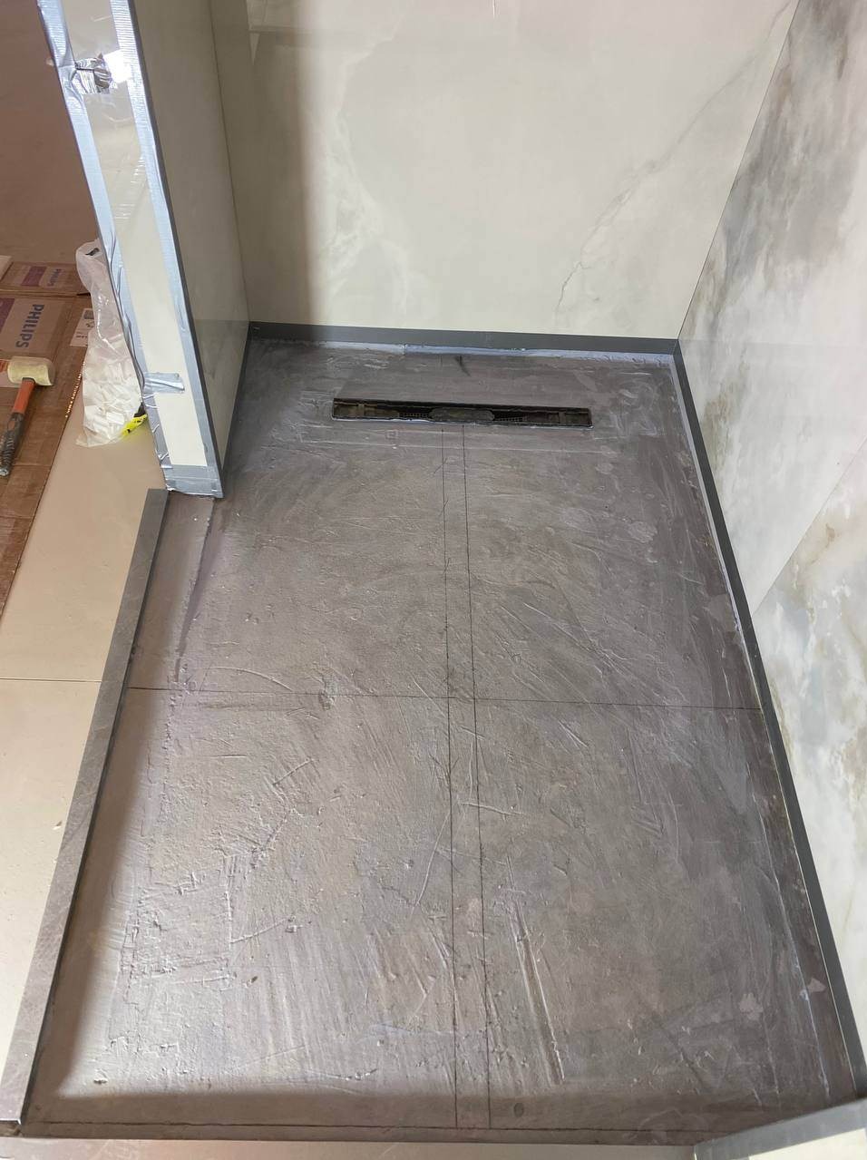 Ukończony brodzik z odpływem liniowym w łazience, przygotowany do położenia płytek. Widoczne linie podziału. Ściany wykończone płytkami imitującymi marmur.