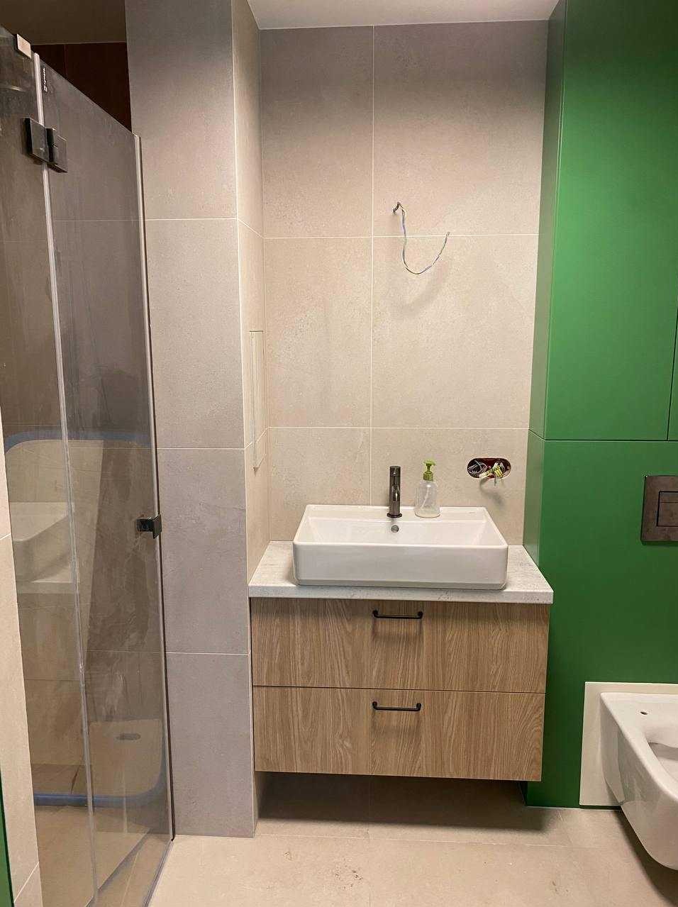 Nowoczesna łazienka z kabiną prysznicową, umywalką na szafce z drewnianym frontem i zielonymi ścianami. Widoczne elementy instalacji elektrycznej przed wykończeniem.