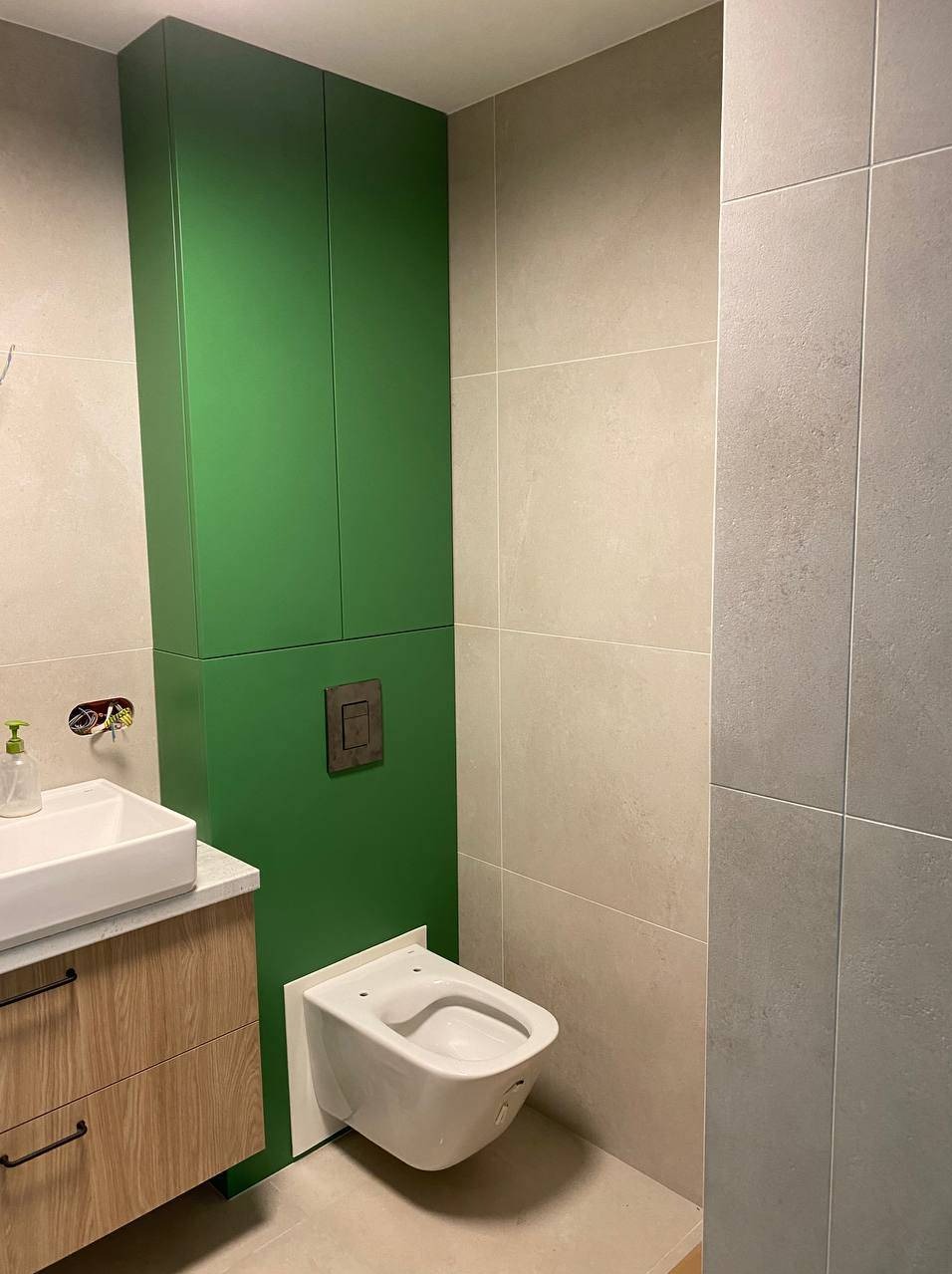 Wykończona łazienka z wiszącą toaletą, umywalką na szafce z drewnopodobnym frontem i zielonymi szafkami, wyłożona płytkami w odcieniach szarości i beżu.