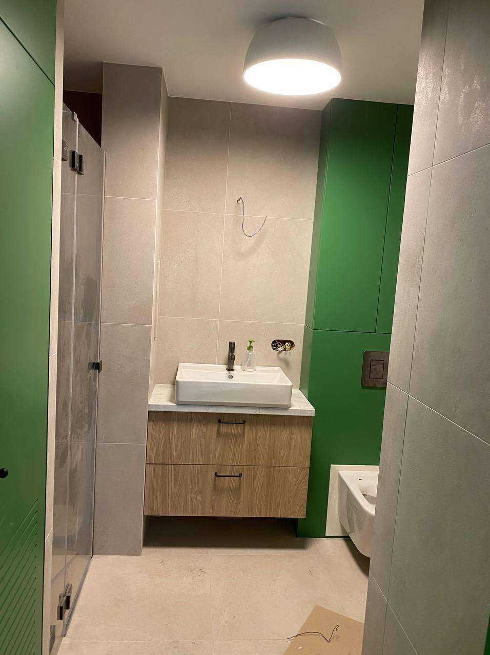 Wykończona łazienka z beżowymi płytkami, drewnianą szafką pod umywalką i zieloną szafą w tle. Widoczna biała umywalka i toaleta.