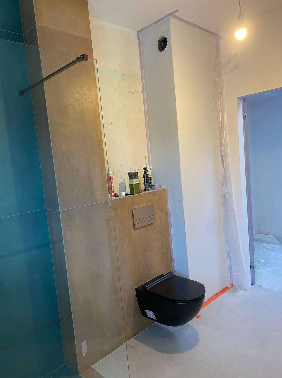 Częściowo wykończona łazienka z zamontowaną czarną toaletą podtynkową, ścianą z beżowych płytek, niebieską kabiną prysznicową i zabezpieczonym obszarem roboczym.