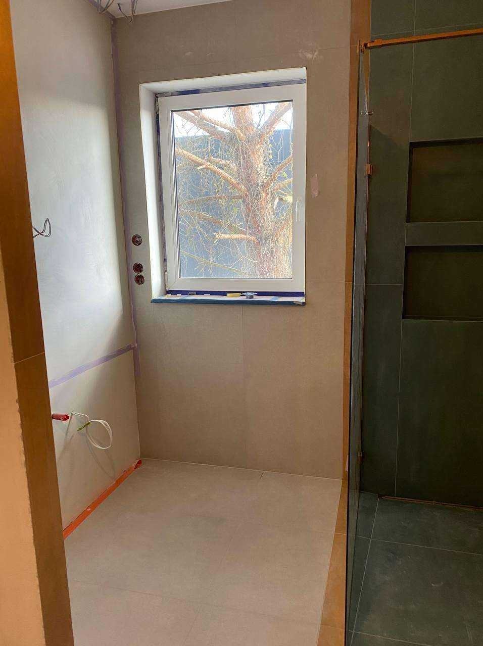 Jasna łazienka w trakcie wykańczania: szare płytki, okno z widokiem na drzewo, instalacje w ścianach, kabina prysznicowa z ciemnymi płytkami.