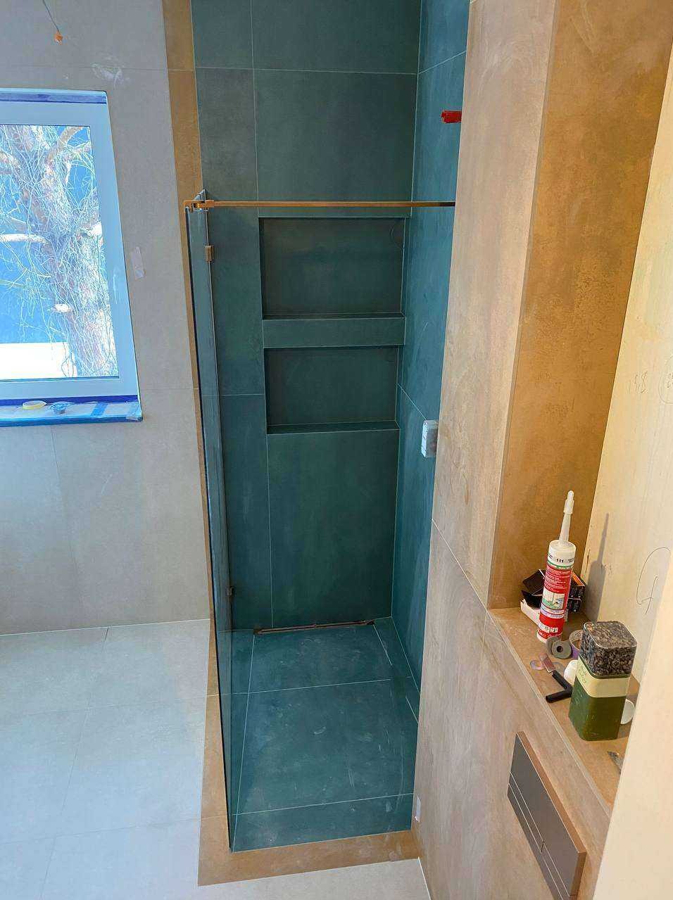 Wykończona kabina prysznicowa z wnękami w ciemnej glazurze, widoczna z boku. Na półce obok narzędzia glazurnicze. Okno z widokiem na drzewo.