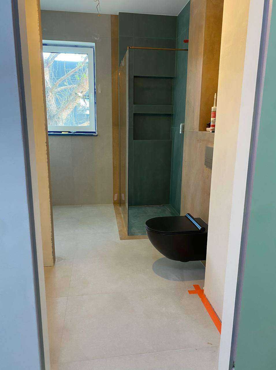 Nowoczesna łazienka z czarną, podwieszaną toaletą i kabiną prysznicową z wnękami. Jasne płytki podłogowe i szare ściany. Widok zza framugi drzwi.