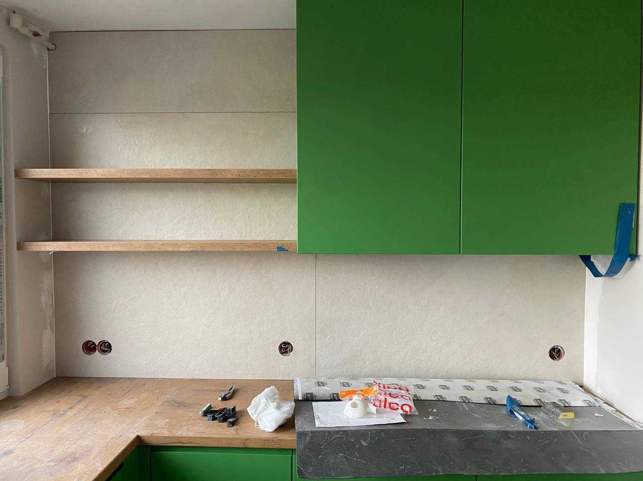 Jasne płytki ścienne z półkami i zielonymi szafkami kuchennymi. Widoczne narzędzia i akcesoria montażowe na blacie. Gniazdka elektryczne w ścianie.