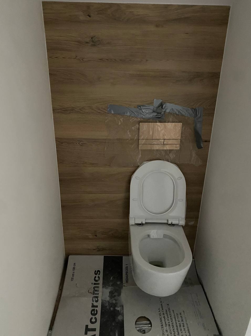 Biała toaleta wisząca na tle ściany wyłożonej panelami imitującymi drewno. Widoczne zabezpieczenia i kartony płytek na podłodze.