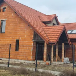 Surowy dom z czerwonej cegły z oknami i dachem z dachówki, otoczony siatką. Widoczne elementy konstrukcyjne i fragment krajobrazu.