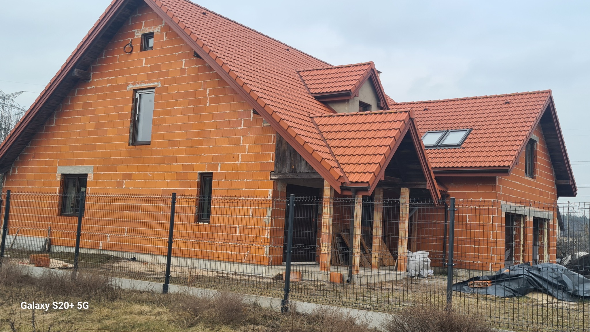 Surowy dom z czerwonej cegły z oknami i dachem z dachówki, otoczony siatką. Widoczne elementy konstrukcyjne i fragment krajobrazu.