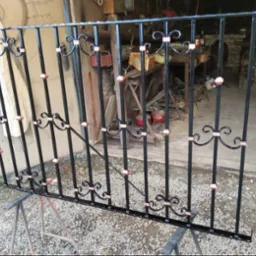 Czarno-miedziana, dekoracyjna balustrada metalowa, ustawiona na kozłach w warsztacie, z widocznymi elementami kutymi i surowym tłem.