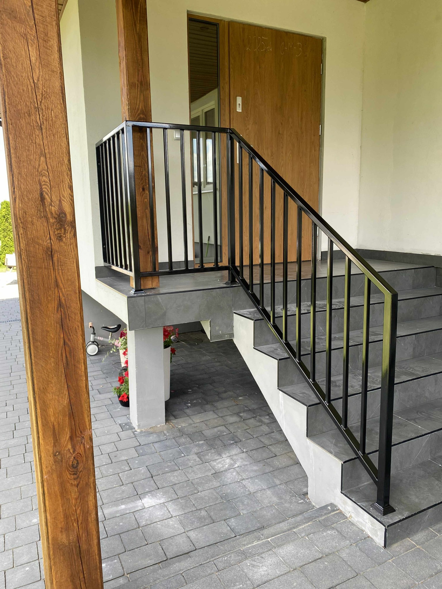 Nowoczesna, czarna balustrada schodowa z metalu z drewnianym słupem. Schody wyłożone szarą płytką, widoczne kwiaty w donicach i dziecięcy rowerek biegowy.