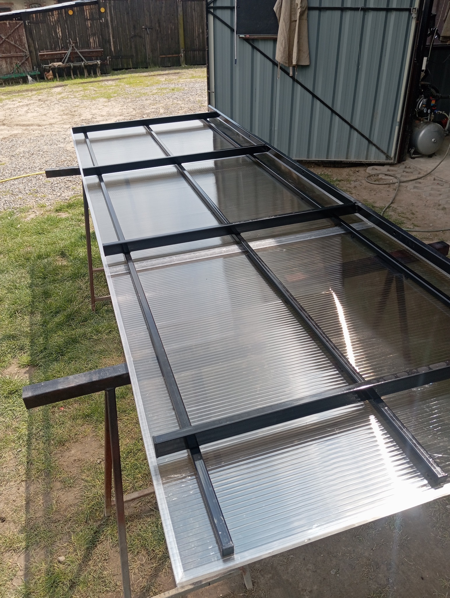 Metalowa konstrukcja z poliwęglanem na kozłach ślusarskich, przygotowana do montażu. Widoczne spawane łączenia i transparentne wypełnienie. Tło: budynek gospodarczy.