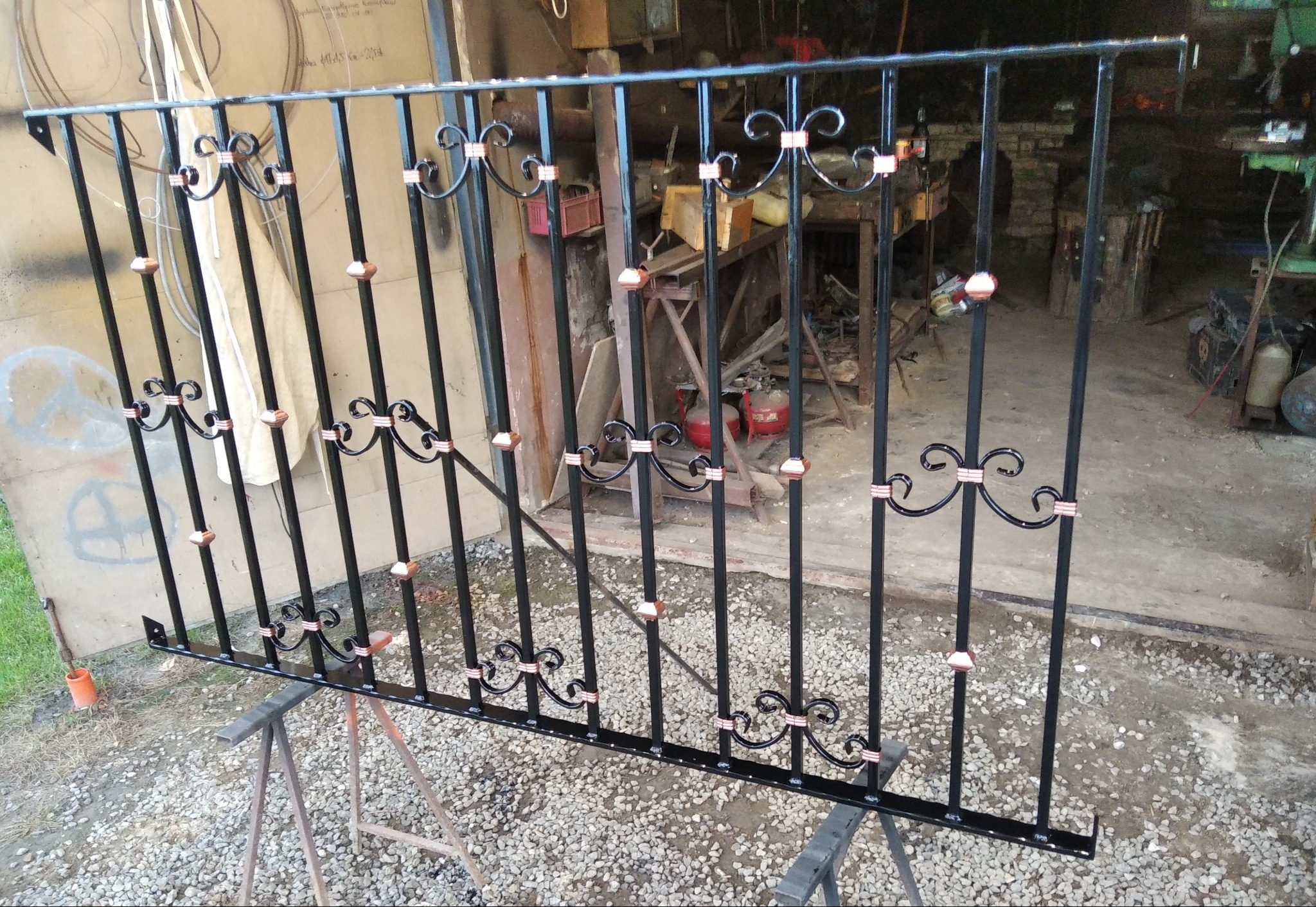 Czarno-miedziana, dekoracyjna balustrada metalowa, ustawiona na kozłach w warsztacie, z widocznymi elementami kutymi i surowym tłem.