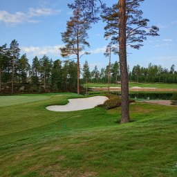 Marek Kolański Ogrody - Pejzaż pola golfowego z piaskowymi bunkrami, zieloną trawą i wysokimi drzewami iglastymi, odbijającymi się w wodzie. Błękitne niebo z delikatnymi chmurami w tle.