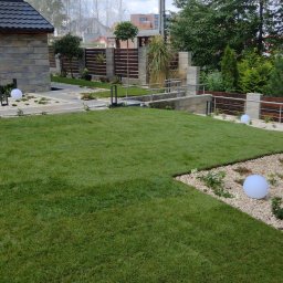 Marek Kolański Ogrody - Zadbany trawnik z rolki, otoczony białym żwirem i kulistymi lampami. Widoczny fragment domu z kamienną elewacją i nowoczesne ogrodzenie z drewnianymi elementami.