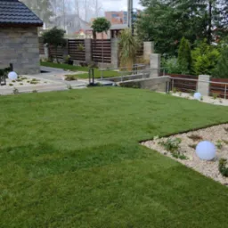 Zadbany trawnik z rolki, otoczony białym żwirem i kulistymi lampami. Widoczny fragment domu z kamienną elewacją i nowoczesne ogrodzenie z drewnianymi elementami.