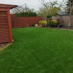 Marek Kolański Ogrody - Zadbany trawnik z widocznymi śladami po aeracji, otoczony ozdobnymi krzewami i elementami architektury ogrodowej, takimi jak drewniana pergola i huśtawka.