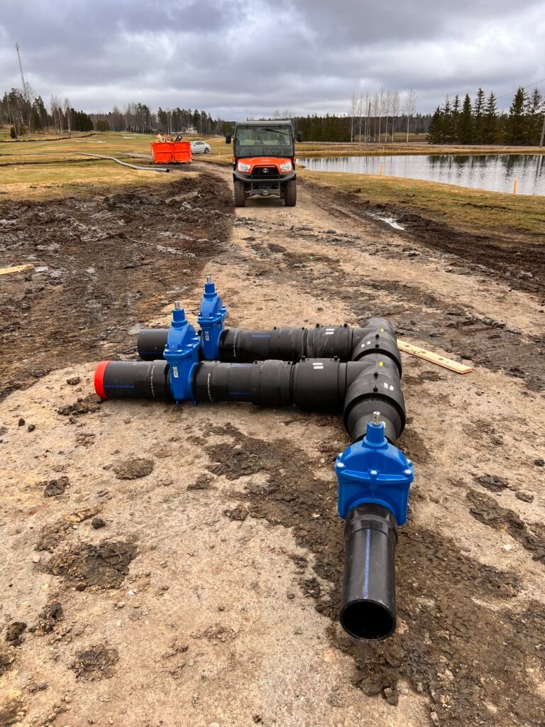 Rozbudowany system rur z niebieskimi zaworami na błotnistym terenie, w tle pojazd Kubota i zbiornik, prawdopodobnie na terenie budowy systemu nawadniania.