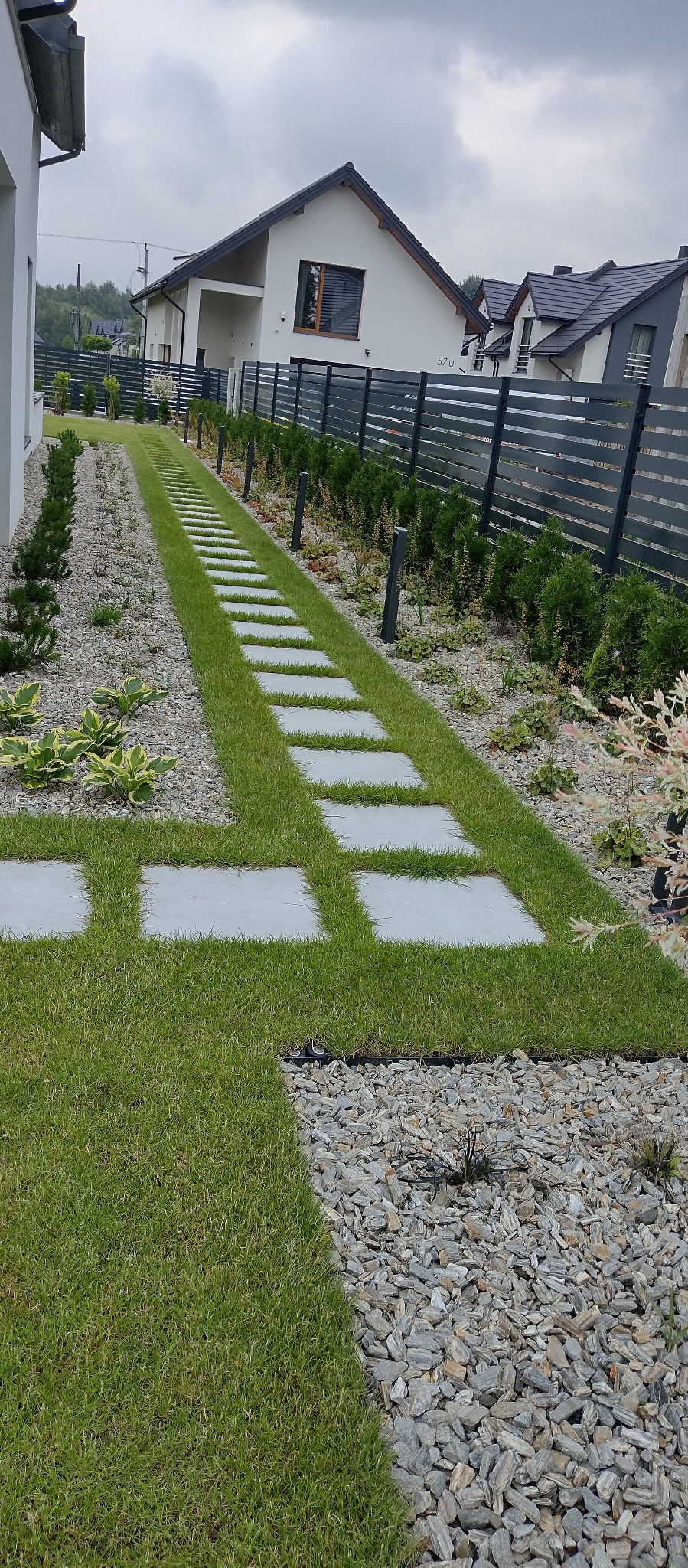 Ogród z równo przystrzyżonym trawnikiem i ścieżką z płyt, otoczony kamieniami i niskimi krzewami. W tle dom z szarą elewacją i ogrodzenie.
