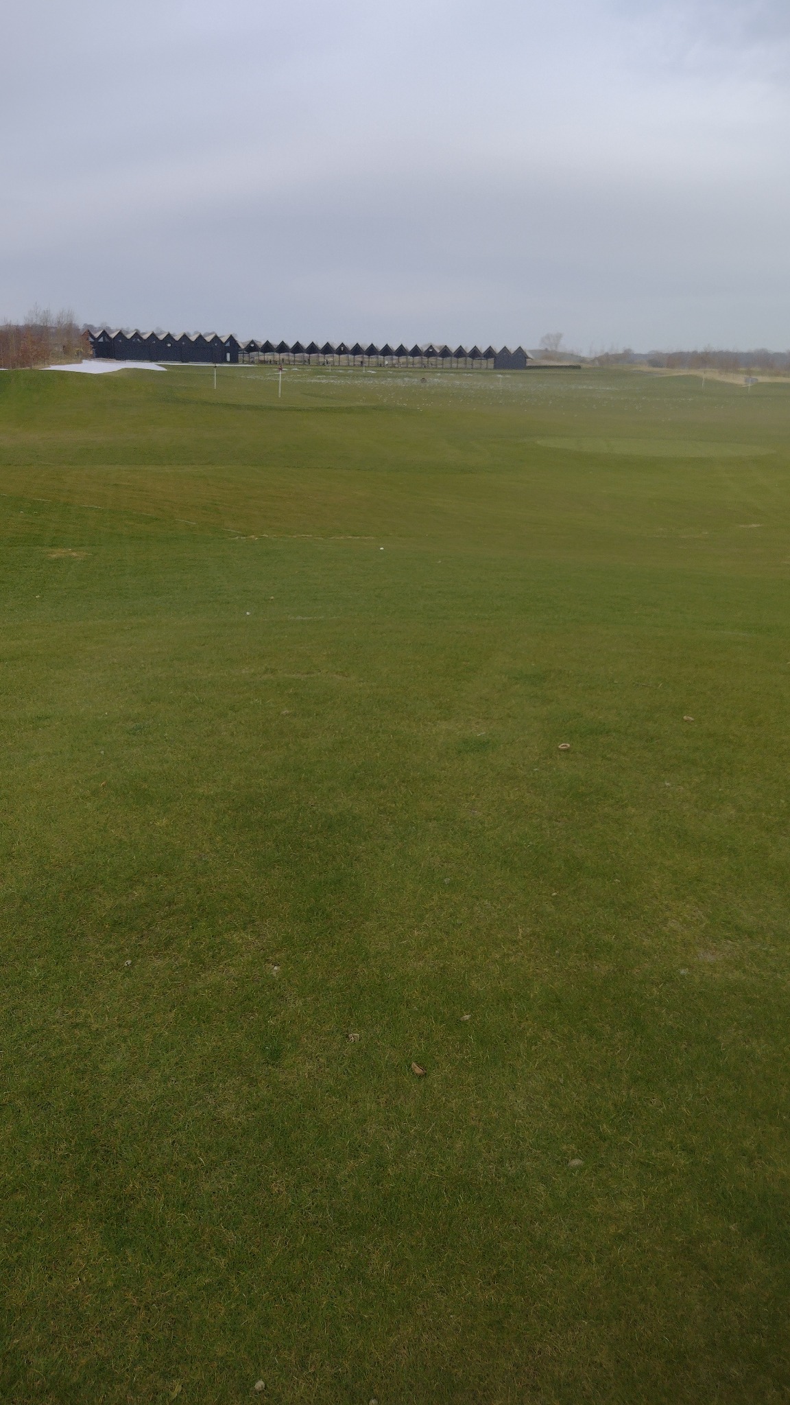 Rozległy trawnik golfowy z rzędem charakterystycznych, ciemnych budek na horyzoncie. Częściowo pokryty śniegiem, pochmurne niebo.