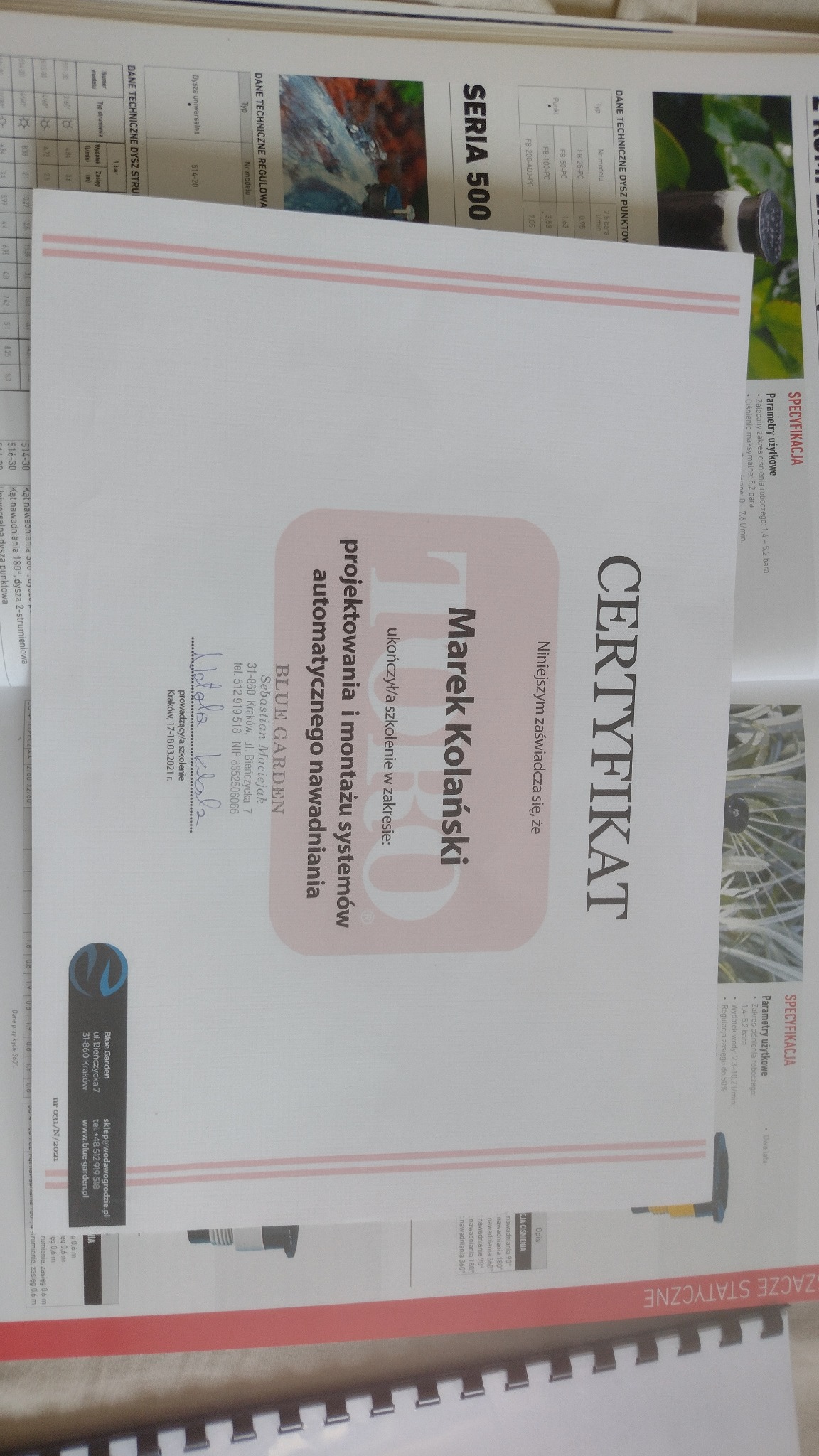Certyfikat ukończenia szkolenia z projektowania i montażu systemów automatycznego nawadniania, wydany przez Blue Garden, prezentowany na tle katalogu.