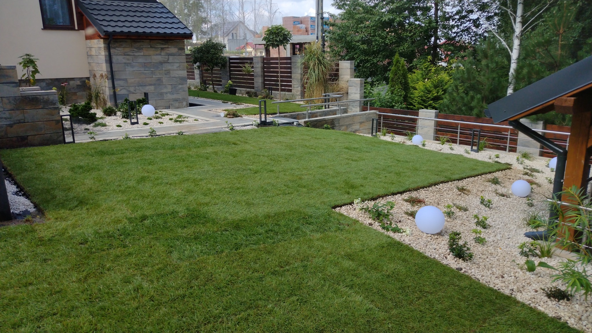Zadbany trawnik z rolki, otoczony białym żwirem i kulistymi lampami. Widoczny fragment domu z kamienną elewacją i nowoczesne ogrodzenie z drewnianymi elementami.