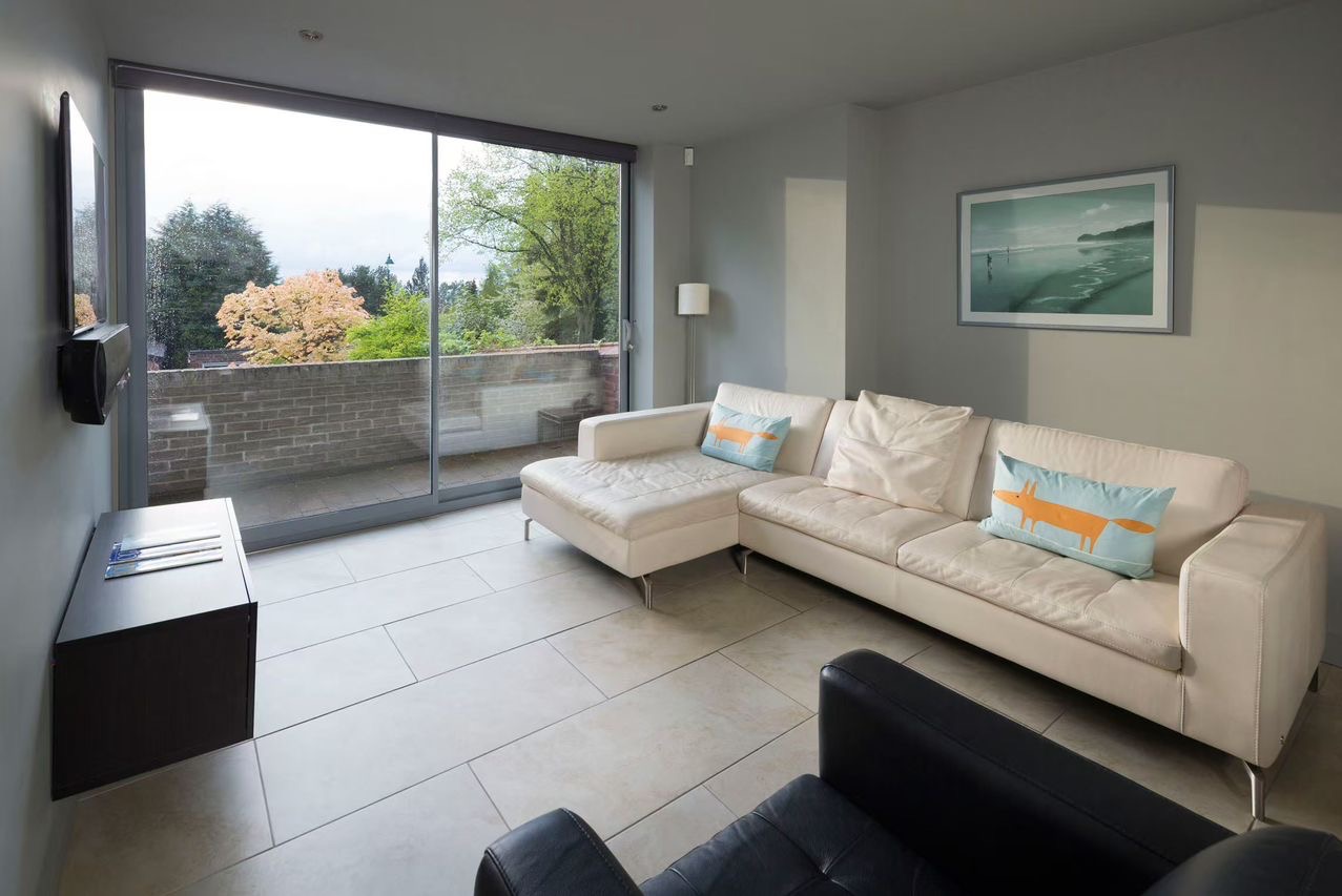 Salon z dużym, przesuwnym oknem balkonowym z widokiem na zieleń. Szklane drzwi balkonowe w szarej ramie wpuszczają dużo światła.