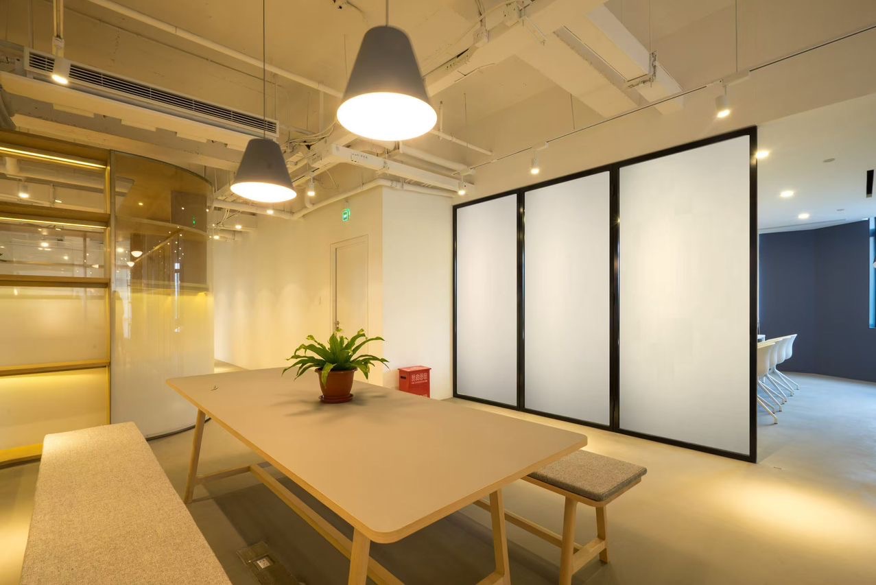 Nowoczesne biuro z szklanymi ścianami działowymi, jasnym stołem i designerskimi lampami wiszącymi. Minimalistyczny wystrój, roślina doniczkowa na stole.