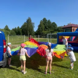 Dzieci bawią się kolorową chustą animacyjną na trawie, w tle dmuchaniec i animator w czerwonej sukience. Słoneczny dzień, błękitne niebo.