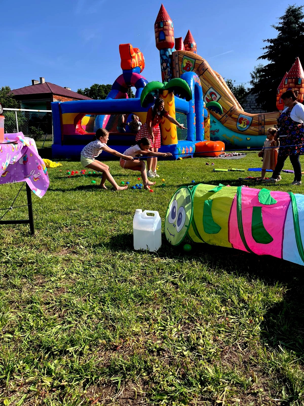 Dzieci rywalizują w limbo na trawie, kolorowy tunel i dmuchany zamek w tle. Zabawa na świeżym powietrzu podczas imprezy dla dzieci.