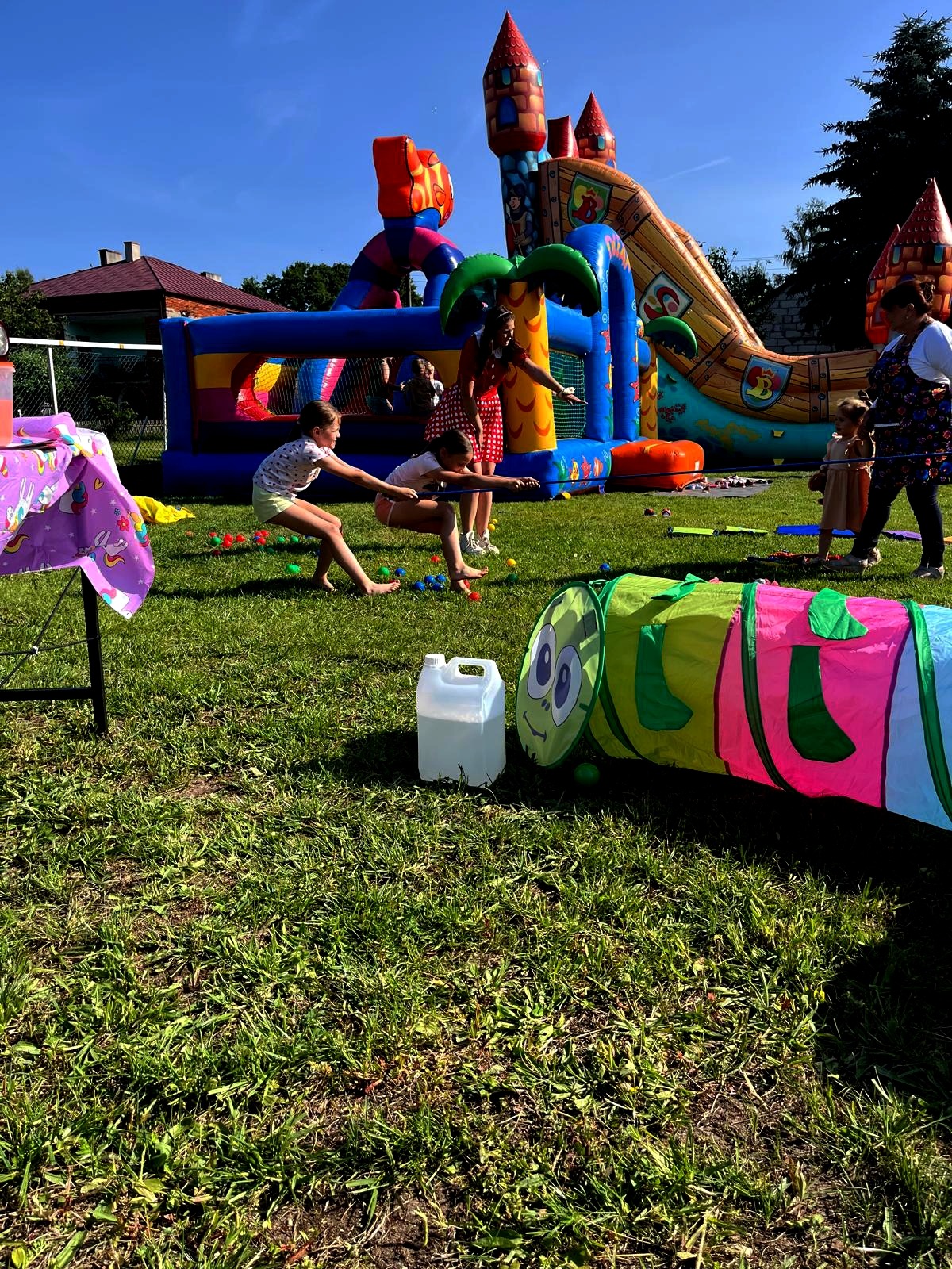 Dzieci bawią się na trawie podczas imprezy plenerowej. W tle dmuchane zamki i zjeżdżalnia. Animatorzy prowadzą zabawy z liną. Kolorowy tunel dla dzieci na pierwszym planie.