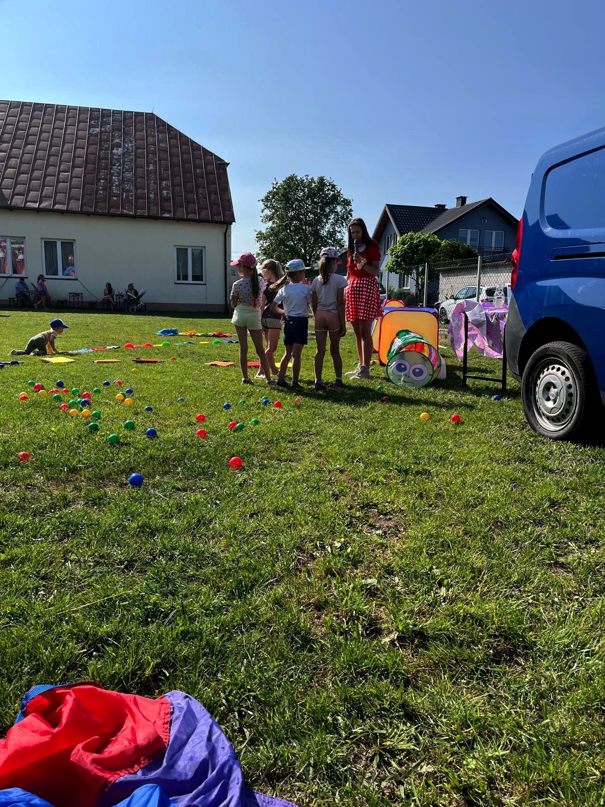 Animator w czerwonej sukience prowadzi gry i zabawy dla grupy dzieci na trawie, kolorowe piłki i tunel dla dzieci, w tle budynki i niebieski van.