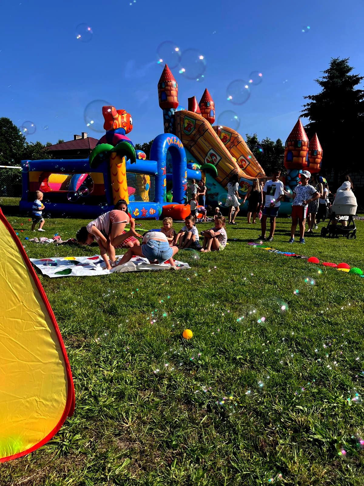 Dzieci bawią się na trawie podczas imprezy plenerowej z dmuchanym zamkiem, tworząc radosną atmosferę pełną śmiechu i zabawy w słoneczny dzień.